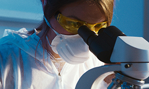 woman in white coat and mask looking into a microscope