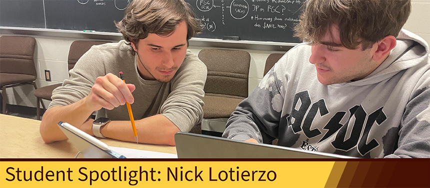 A photograph of Nick Lotierzo tutoring a student. Both men are seated at a blonde wooden table in the CS drop-in tutoring room. There is a blackboard with various equations behind them, as well as brown, felt chairs lined up in a row against the beige wall. Nick is a young white man, wearing a long sleeved gray shirt pushed up to his elbows. He has brown hair swept to either side of his face. He has a thin brown beard. He’s pointing with a pencil at a tablet screen. The tablet is blue, with an electronic pen resting on top. The tutee is another white young man, with a partial beard and dirty blonde hair. He is wearing a gray AC/DC sweatshirt. He is looking to where Nick is pointing. On the table, you can see the top quarter of his silver laptop.   At the bottom of the screen is a graphic. The graphic is rectangular, with its border Rowan brown and it’s color Rowan yellow. At the right fourth of the page, the yellow fades into various shades of Rowan brown. The left, yellow part of the graphic reads, “Student Spotlight: Nick Lotierzo.” 