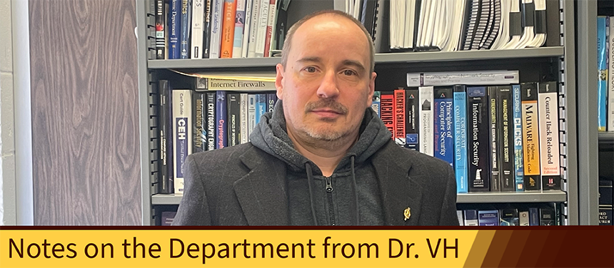 A photograph depicting Dr. Vasil Hnatyshin standing in front of the bookcase in his office. Dr. Hnatyshin is a white man with salt-and-pepper hair and a partial beard. He is staring at the camera, not smiling. He is wearing a gray sweatjacket with a black suit jacket on top. On the bottom left corner of the photo, a Rowan yellow rectangle extends across the bottom sixth of the photograph, fading into a Rowan brown gradient. The rectangle has text on it that reads, "Notes on the Department from Dr. VH."