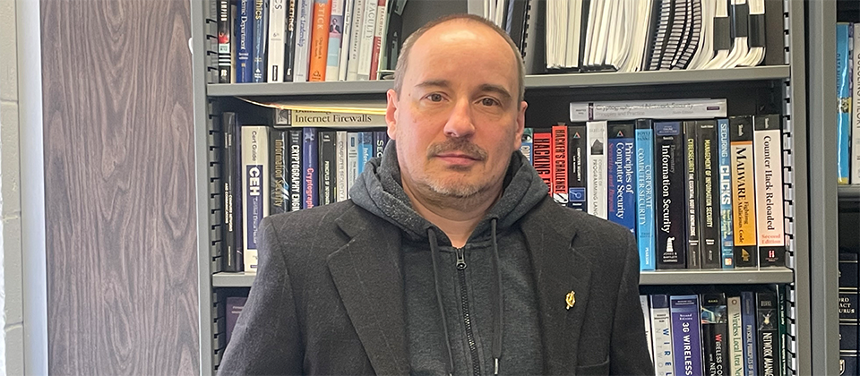 A photograph depicting Dr. Vasil Hnatyshin standing in front of the bookcase in his office. Dr. Hnatyshin is a white man with salt-and-pepper hair and a partial beard. He is staring at the camera, not smiling. He is wearing a gray sweatjacket with a black suit jacket on top. 