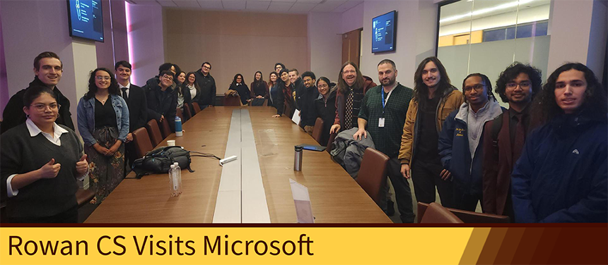 A group photo of many CS students in a Microsoft corporate meeting room. There is an upside down trapezoid hanging on the top part of the image.  On the bottom left corner of the photo, a Rowan yellow rectangle extends across the bottom sixth of the photograph, fading into a Rowan brown gradient. The rectangle has text on it which reads, "Rowan CS Takes Microsoft."