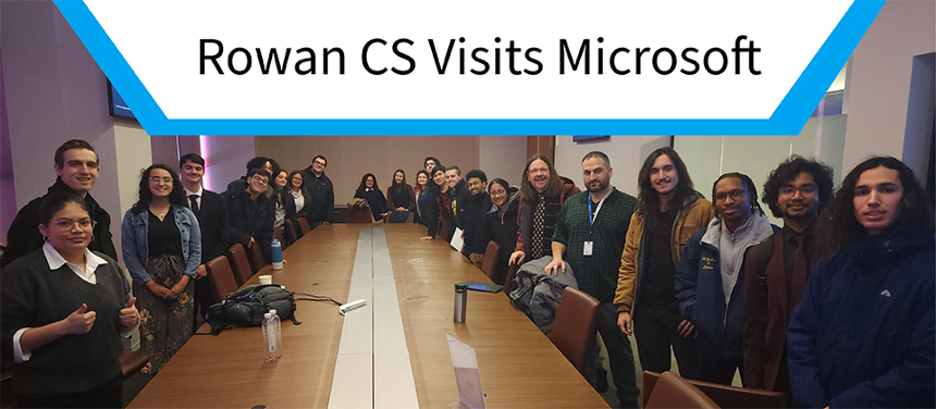 A group photo of many CS students in a Microsoft coporate meeting room. There is an upside down trapezoid hanging on the top part of the image. The trapezoid is white with a  bright blue border. The trapezoid has text inside which reads, "Rowan CS Visits Microsoft".