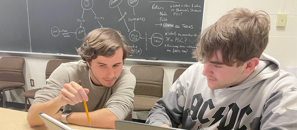 A photograph of Nick Lotierzo tutoring a student. Both men are seated at a blonde wooden table in the CS drop-in tutoring room. There is a blackboard with various equations behind them, as well as brown, felt chairs lined up in a row against the beige wall. Nick is a young white man, wearing a long sleeved gray shirt pushed up to his elbows. He has brown hair swept to either side of his face. He has a thin brown beard. He’s pointing with a pencil at a tablet screen. The tablet is blue, with an electronic pen resting on top. The tutee is another white young man, with a partial beard and dirty blonde hair. He is wearing a gray AC/DC sweatshirt. He is looking to where Nick is pointing. On the table, you can see the top quarter of his silver laptop. 